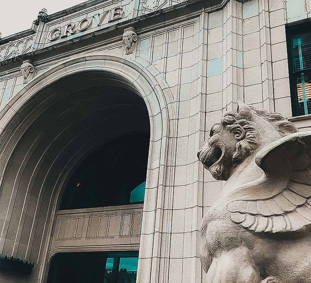 a side angle shot of the Grove Arcade's north entrance, in the foreground is one of the winged lions, the archway and the "Grove" sign are visible in the background.